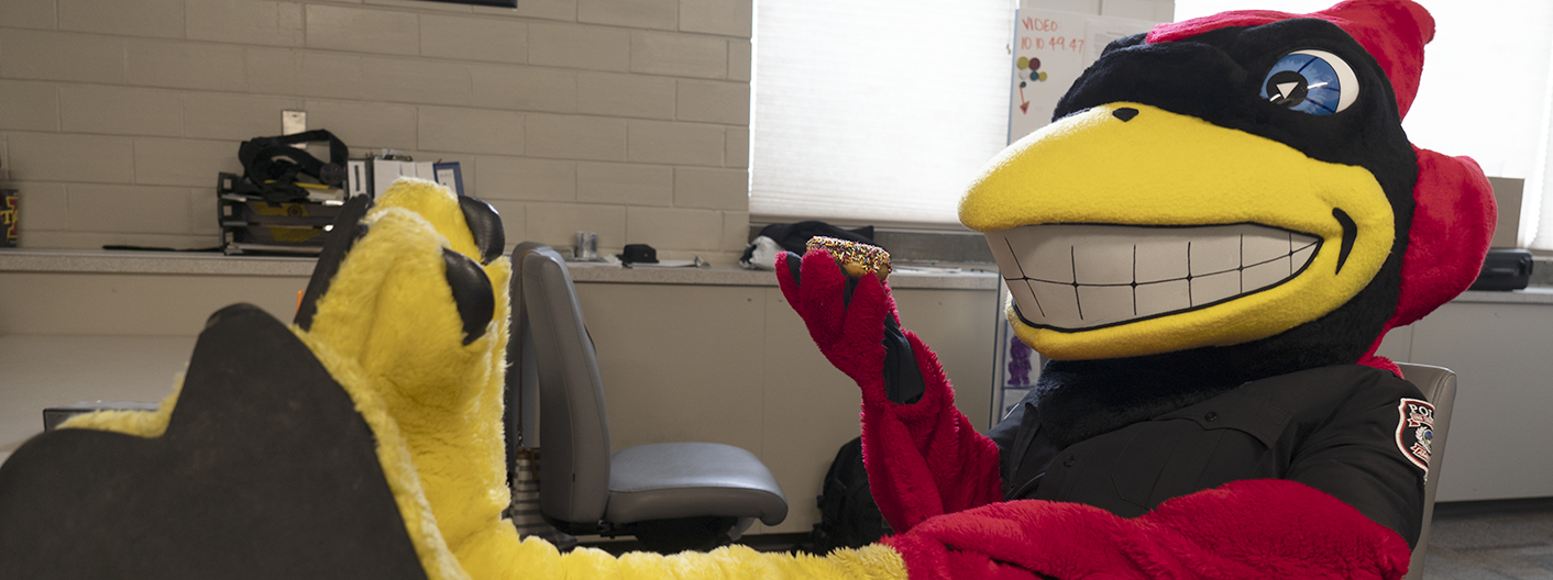 Cy mascot with his feet up on the desk, holding a donut.